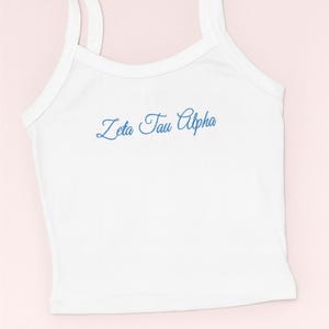 Könnte beinhalten: Weißes Tanktop mit dünnen Trägern und dem gestickten Text "Zeta Tau Alpha" in blauer Schrift. Das Oberteil ist aus einem weichen Material gefertigt und eignet sich für Freizeitkleidung. Der Hintergrund ist hellrosa.