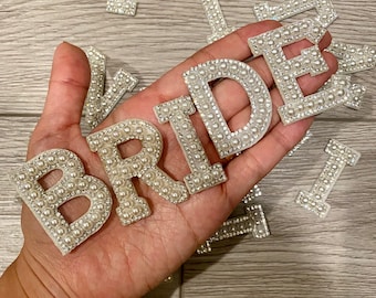 Puede incluir: Letras de fieltro blanco que deletrean "BRIDE" con adornos de perlas y diamantes de imitación.