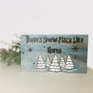 May include: A wooden sign with a blue background and white snowflakes. The sign says "There's Snow Place Like Home" with a black font. There are five white trees in the bottom row of the sign.