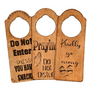 May include: Three light brown wooden door hangers with laser-engraved text and graphics. The first reads Do Not Enter Unless You Have Snacks. The second says Prayin Do Not Disturb with praying hands. The third says Kindly go away with sleepy eyes.