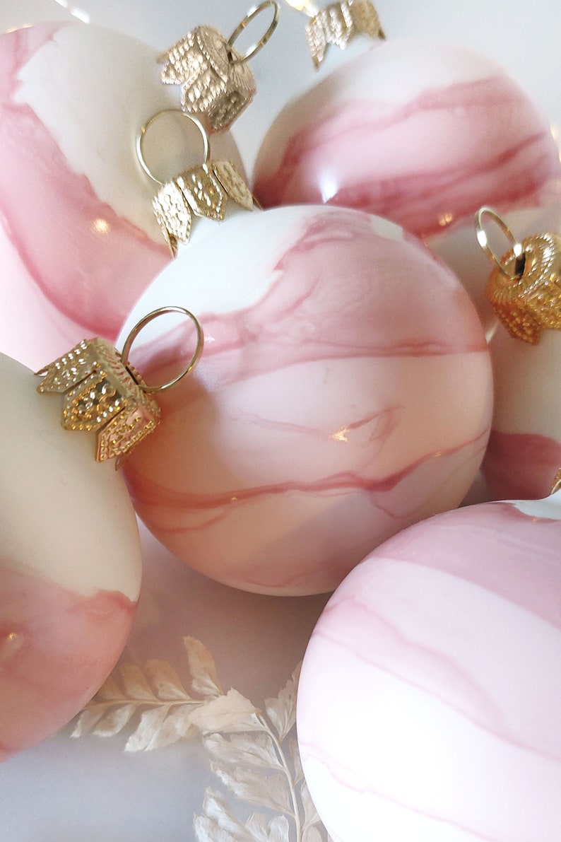 May include: Four pink and white marble Christmas ornaments with gold accents. The ornaments are round and have a swirl pattern. The ornaments are on a white surface with dried white flowers.