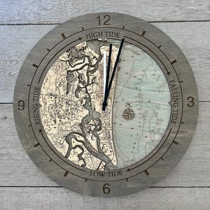 May include: A round wooden clock with a detailed map design. The clock face features a map with tide indicators: Rising Tide, High Tide, Falling Tide, and Low Tide. The clock hands are black, and the frame is gray.