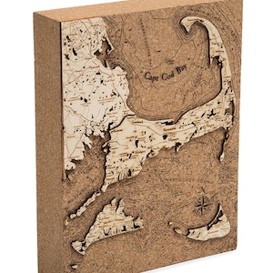May include: A corkboard map of Cape Cod, Massachusetts, with a compass rose in the lower right corner. The map shows the coastline, towns, and bodies of water. The map is made of cork and has a 3D effect.
