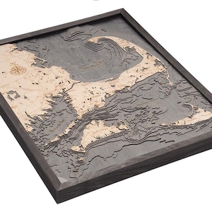 May include: A wooden map of the Cape Cod coastline, framed in a dark gray frame. The map is detailed and shows the various inlets and bays of the area. The map is made of wood and has a 3D effect.
