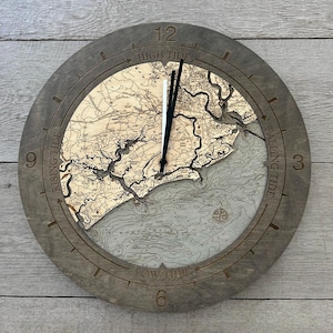 May include: A round wooden clock with a detailed map design. The clock face features a topographic map with coastlines and water depth lines. The clock hands are black, and the clock is labeled with "High Tide", "Falling Tide", "Low Tide", and "Rising Tide".