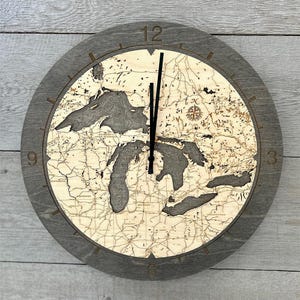 May include: A round wooden wall clock with a gray frame. The clock face features a detailed map of the Great Lakes, with dark gray and beige tones. The clock hands are black.
