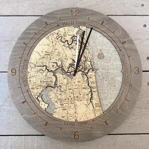 May include: A circular wooden wall clock featuring a detailed nautical map of Jacksonville, Florida. The frame displays the numbers 3, 6, 9, and 12, with text indicating tide phases: High Tide, Falling Tide, Low Tide, and Rising Tide.