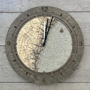 May include: A round wooden clock with a map design. The clock face features a detailed nautical chart and the words "High Tide", "Falling Tide", "Low Tide", and "Rising Tide". The clock hands are black, and the frame is gray.