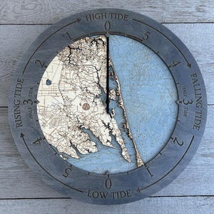 May include: A gray wooden clock with a nautical map design. The clock face features a tide chart with markings for high tide, low tide, rising tide, and falling tide. The clock hands are black.