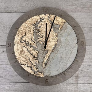 May include: A round wooden wall clock with a detailed map design. The clock face features a topographic map in shades of beige and gray, with black clock hands and a gray wooden frame. The clock measures approximately 12 inches in diameter.