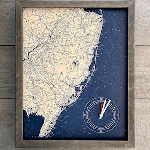 May include: A framed nautical map clock featuring a detailed map of the New Jersey coastline with a tide clock inset. The clock face has a red hour hand and a white minute hand. The tide clock has markings for high tide, low tide, and falling tide.