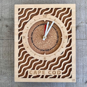 May include: A wooden clock with a cork face and a wave pattern around the edge. The clock face has a tide chart with the words "Rising Tide", "High Tide", "Low Tide", and "Falling Tide". The clock also has the words "Cape Cod" and "Tidal Shoals" printed on it.