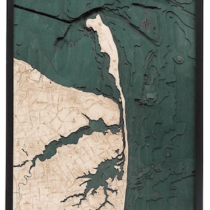 May include: A framed wooden map with a detailed topographical design. The map features a coastal area with a dark green ocean and a light wood-colored landmass. The frame is black, and the map has a three-dimensional appearance.