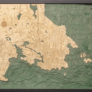 May include: A wooden map of Victoria, British Columbia, with a green and brown color scheme. The map shows the city's streets, waterways, and surrounding areas. The map is framed in a black frame.