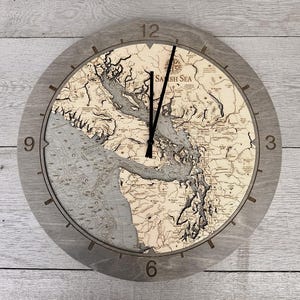 May include: A round wooden wall clock with a detailed map of the Salish Sea. The clock face features a light wood grain design with black clock hands and a gray wooden frame. The numbers 3, 6, 9, and 12 are visible.