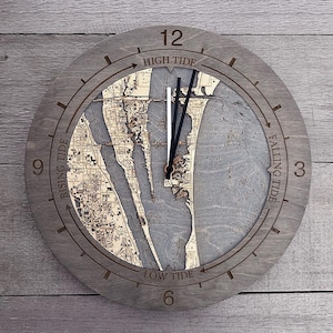 May include: A round, gray wooden clock with a detailed map design in the center. The clock face features the words "High Tide", "Falling Tide", "Low Tide", and "Rising Tide". The clock hands are black.
