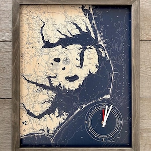 May include: A framed nautical map of the Outer Banks, North Carolina, with a tide clock in the lower right corner. The map is printed on a blue background with white lines and text. The tide clock has a white face with black markings and red and white hands.