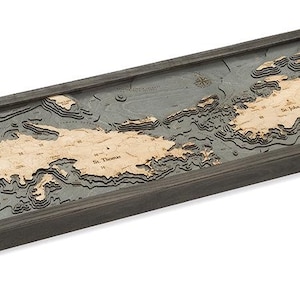 May include: A wooden 3D map of the Virgin Islands, with a dark gray frame. The map is detailed and shows the islands' topography, with different shades of brown and gray representing different elevations. The map is mounted on a wall.