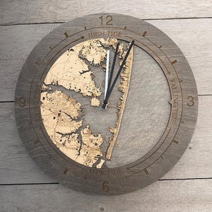 May include: A round, gray wooden clock with a detailed map design. The clock face features a map of a coastal area, with the time indicated by black hands. The words "High Tide", "Falling Tide", "Low Tide", and "Rising Tide" are engraved on the clock face.
