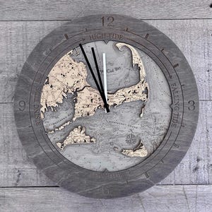 May include: A round, gray wooden clock with a detailed map of Cape Cod and surrounding waters. The clock face features black hands, labeled "High Tide," "Falling Tide," "Low Tide," and "Rising Tide." The map is in shades of beige and gray.