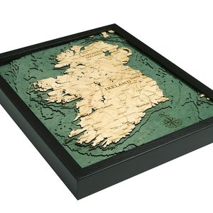 May include: A framed 3D wood map of Ireland with a green and brown color scheme. The map is mounted in a black frame and includes a compass rose in the lower right corner.