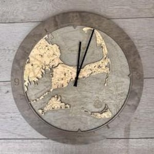 May include: Round wooden clock featuring a detailed map of Cape Cod. The clock face is light brown with a gray border and black clock hands. The numbers 9, 12, 3, and 6 are visible. The clock measures approximately 12 inches in diameter.