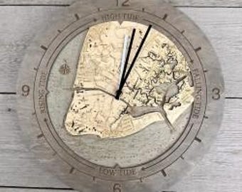 May include: A round wooden wall clock featuring a light-colored wood map inlay of a coastal area. The clock face has engraved text reading High Tide, Rising Tide, Falling Tide, and Low Tide, with black and silver hands and a compass rose icon.