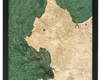 May include: A framed wooden map of the Monterey Peninsula, featuring detailed topographical lines and place names. The map has a natural wood color for the land and a teal-green hue for the water, with a compass rose at the bottom. The frame is black.