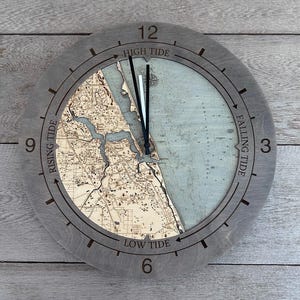 May include: A gray wooden clock with a map of a coastal area in the center. The clock face has Roman numerals and the words "High Tide", "Low Tide", "Rising Tide", and "Falling Tide" around the edge.