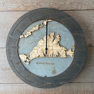 May include: A wooden clock with a nautical map of Martha's Vineyard. The clock face has a compass rose and tide markings. The clock hands are black and the clock face is gray with a light blue background. The clock is framed in dark gray wood.