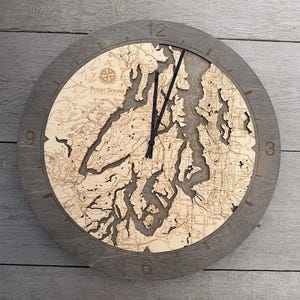 May include: A round wooden wall clock with a gray frame. The clock face features a detailed map of Puget Sound, with laser-cut waterways and landmasses. Black clock hands indicate the time. The clock is a decorative home decor item.