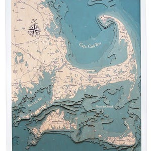 May include: A framed 3D wooden map of Cape Cod, Massachusetts, with detailed topographical features. The map features the text "WOOD CHART" and a compass rose. The water is a teal color, and the land is a light wood color.
