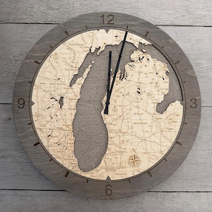 May include: A round wooden wall clock with a gray frame and a detailed map of the Great Lakes region. The clock face features the states of Michigan, Wisconsin, Illinois, and Indiana. Black clock hands indicate the time. The clock is approximately 12 inches in diameter.