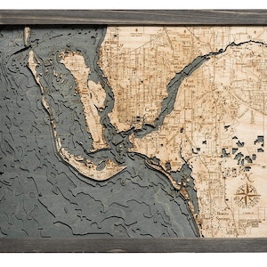 May include: A detailed wooden map of the Fort Myers, Florida area, with a gray background and a compass rose in the lower right corner. The map shows the coastline, waterways, and major roads. The text "Fort Myers" is visible in the upper center of the map.