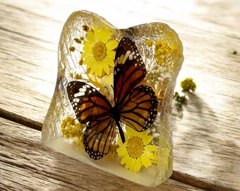 May include: A monarch butterfly encased in clear resin with yellow flowers. The butterfly is brown and black with orange markings. The flowers are pressed and preserved in the resin.