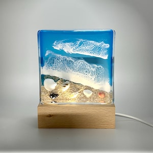 May include: A square resin lamp with a blue ocean scene, white waves, and a sandy beach with seashells. The lamp is on a wooden base and has a white cord.