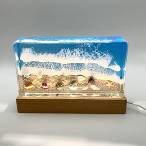May include: A rectangular clear resin lamp with a beach scene inside. The scene includes blue water, white waves, sand, and seashells. The lamp has a wooden base and a cord.