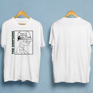 Puede incluir: Camiseta blanca con un gráfico en blanco y negro de Lisa Simpson con el texto "MEAT IS MURDER" en su sombrero. Las palabras "THE SIMPSONS" están impresas verticalmente en verde. La camiseta se muestra en una percha de madera.