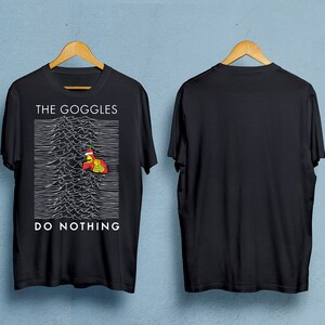 Puede incluir: Camiseta negra con el texto "THE GOGGLES DO NOTHING" encima y debajo de un gráfico blanco. El gráfico presenta un personaje de dibujos animados con un gorro de Papá Noel. La camiseta se muestra en una percha de madera.