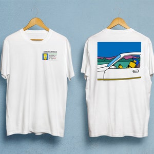 Könnte beinhalten: Weißes T-Shirt mit Cartoon-Grafiken auf Vorder- und Rückseite. Auf der Vorderseite befindet sich ein Führerschein mit der Aufschrift "SPRINGFIELD" und einer Cartoonfigur. Auf der Rückseite ist eine Cartoonfigur zu sehen, die ein weißes Auto fährt.