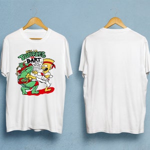 May include: White t-shirt featuring a colorful graphic of Bart Simpson in a karate stance facing a Teenage Mutant Ninja Turtle. The text "Ninja Turtles vs Bart" is above the characters. The shirt is displayed on a wooden hanger.