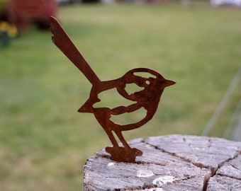 Könnte beinhalten: Rostige Metallvogel-Silhouette, die auf einem verwitterten Baumstumpf sitzt. Die Form des Vogels ist ausgeschnitten und gibt den Hintergrund frei. Die Skulptur hat einen langen Schwanz und steht vor einem verschwommenen grünen Hintergrund.