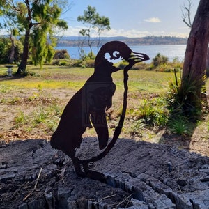 Puede incluir: Silueta de pingüino de metal negro de pie sobre un tocón de árbol. El pingüino está mirando hacia el lado derecho de la imagen. El fondo es una imagen borrosa de un lago y árboles.