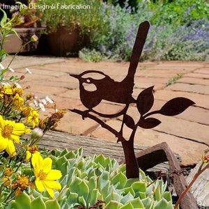 Puede incluir: Una silueta de metal oxidado de un pájaro posado en una rama, con hojas, sobre un fondo de plantas verdes y adoquines de ladrillo marrón.