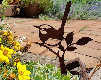 Könnte beinhalten: Eine rostige Metallsilhouette eines Vogels, der auf einem Ast mit Blättern sitzt, vor einem Hintergrund aus grünen Pflanzen und braunen Backsteinpflastersteinen.