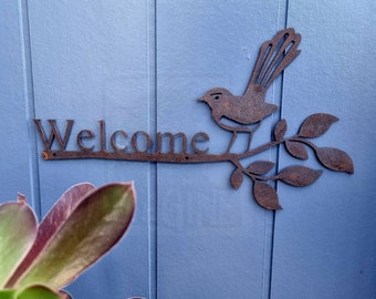 Könnte beinhalten: Ein rostiges Willkommensschild aus Metall mit dem Wort "Welcome" und einem Vogel, der auf einem Zweig mit Blättern sitzt. Das Schild ist an einer blauen Holzoberfläche befestigt.