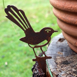 Puede incluir: Una estaca de jardín de metal oxidado con forma de pájaro. El pájaro tiene una cola detallada y está posado sobre una pequeña base. La estaca está junto a una maceta de terracota y un tocón de madera, con un fondo de hierba verde.