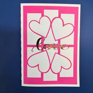 May include: A greeting card with a pink border and a white interior featuring four white heart shapes. The word "love" is written in gold script across the center of the card. The background is a solid dark blue.