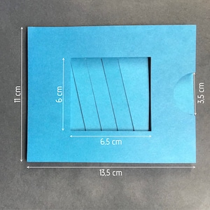 May include: A blue cardstock template for a pop-up card. The template is 13.5 cm wide, 11 cm tall, and has a 6.5 cm by 6 cm opening. The opening has five parallel lines cut into it.