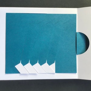 May include: A teal blue card with a white circle cut out on the right side. The left side of the card has five white triangles cut out, creating a layered effect.